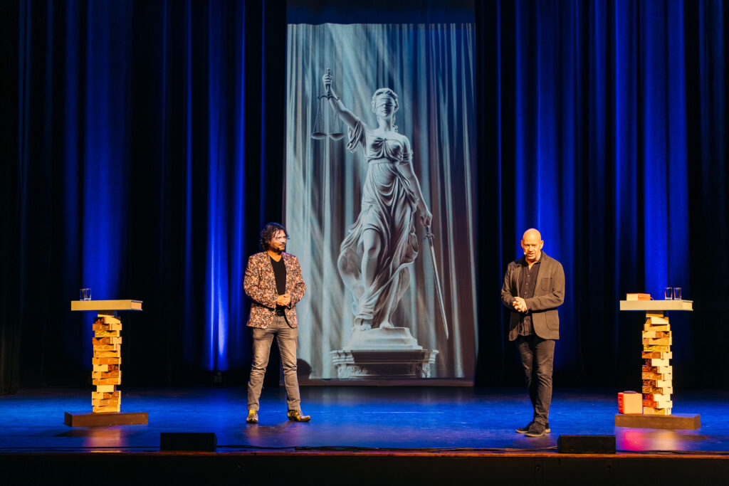 Job Knoester en Dennis Jansen staan op het podium tijdens hun theatercollege over TBS, met achter hen een afbeelding van Vrouwe Justitia.