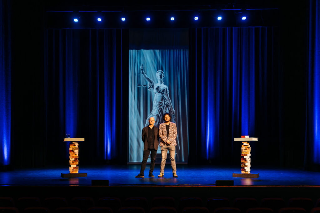Job Knoester en Dennis Jansen op theaterpodium voor TBS-tour, met beeld van Vrouwe Justitia op de achtergrond.