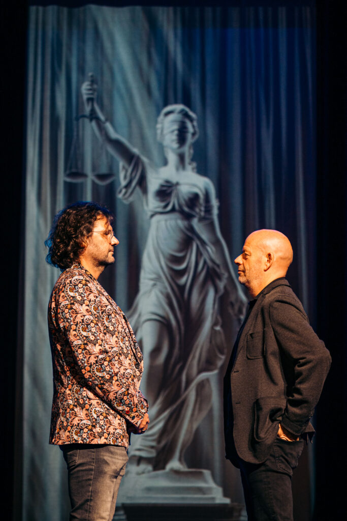 Tbs-advocaat Job Knoester en journalist Dennis Jansen op het podium tijdens theatershow over tbs en strafrecht, met Vrouwe Justitia op de achtergrond.
