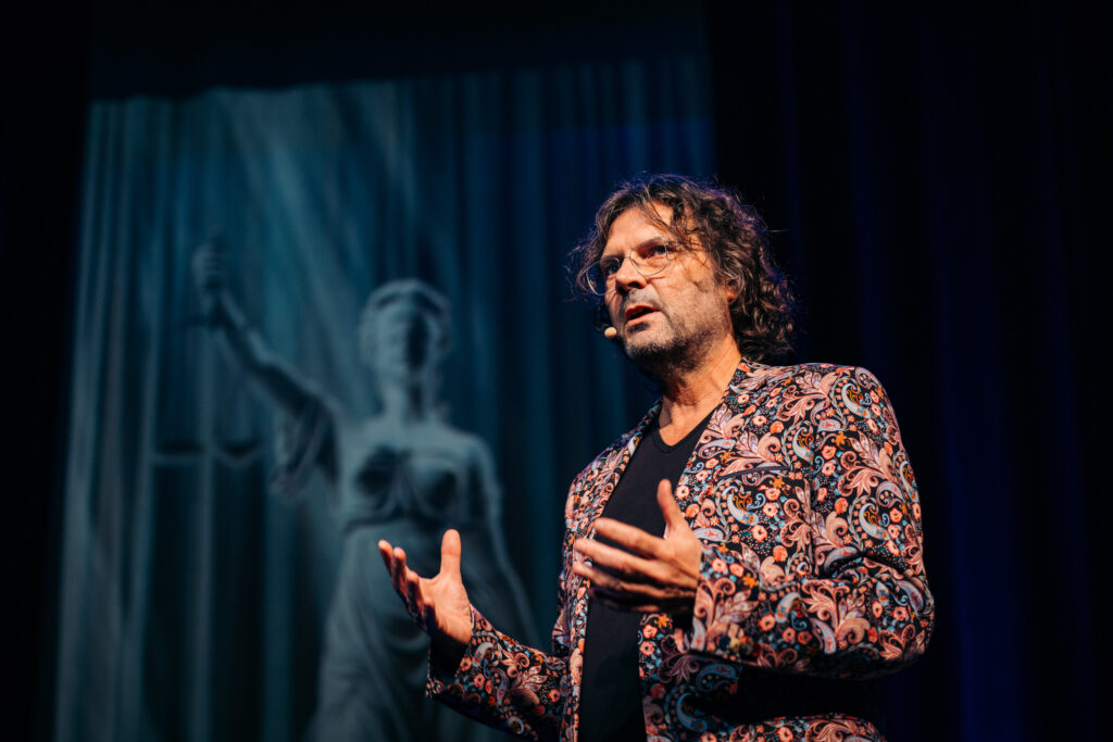 Job Knoester spreekt op een theaterpodium tijdens een voorstelling over tbs, met microfoon en decor op de achtergrond